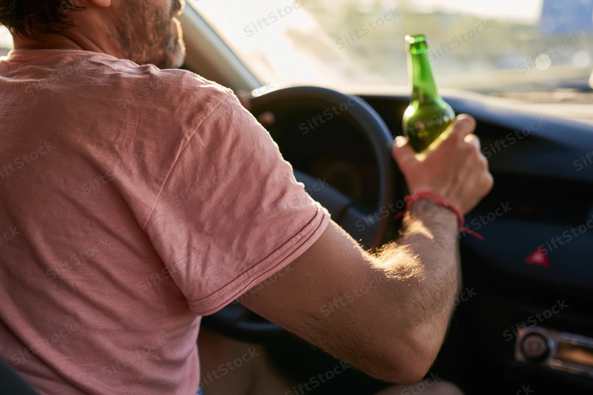 Man driving a car holding a green beer bottle in his hand inside the vehicle