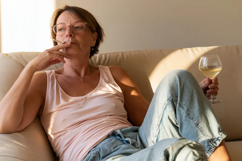 Middle-aged woman reclining on a couch smoking a cigarette and holding a glass of white wine in a sunlit living room