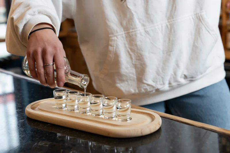 Person pouring alcohol into shot glasses arranged on a wooden tray on a bar countertop