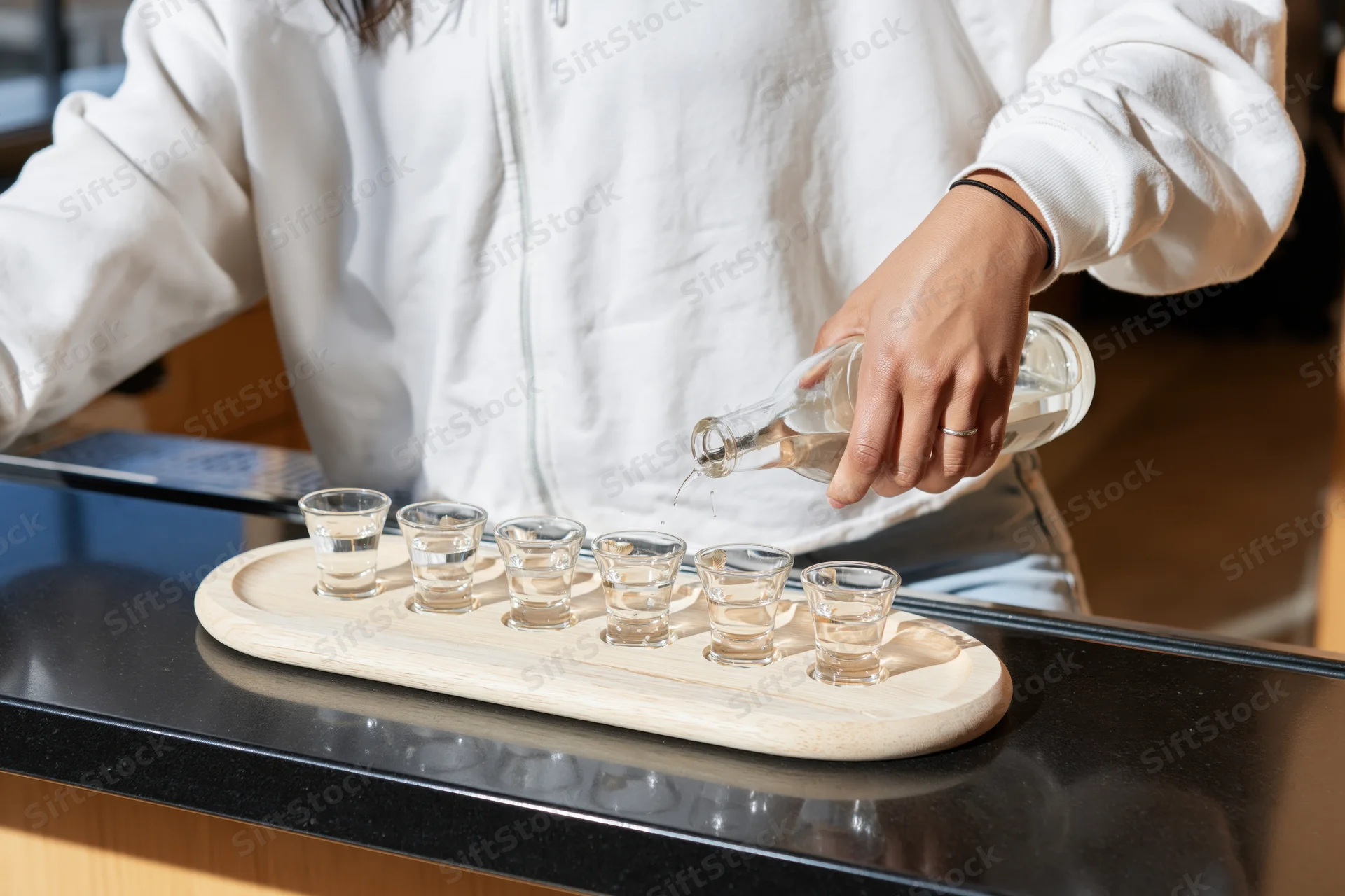 Woman pouring alcohol into shot glasses arranged on a wooden tray on a black countertop