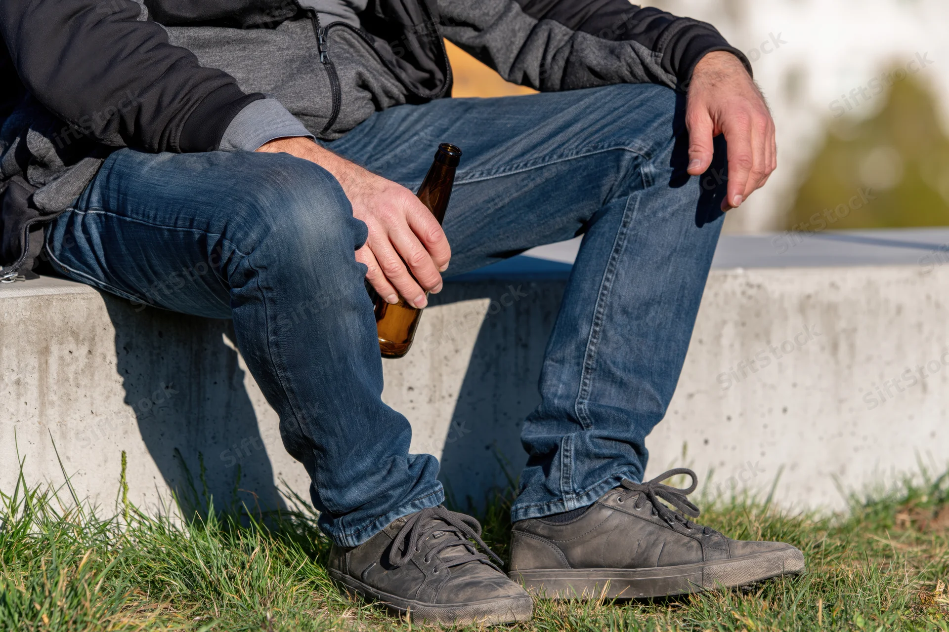 Man sitting on concrete ledge holding beer bottle outdoors on grassy ground