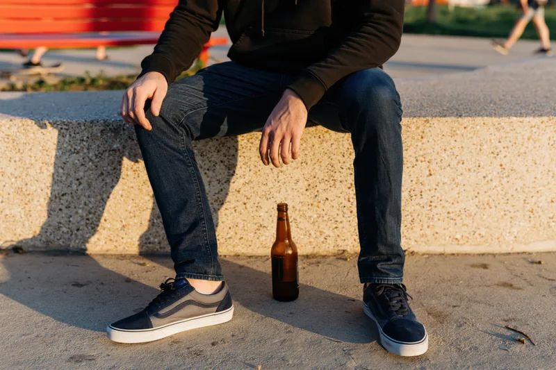 Man sitting on concrete ledge with beer bottle between his feet in urban park setting