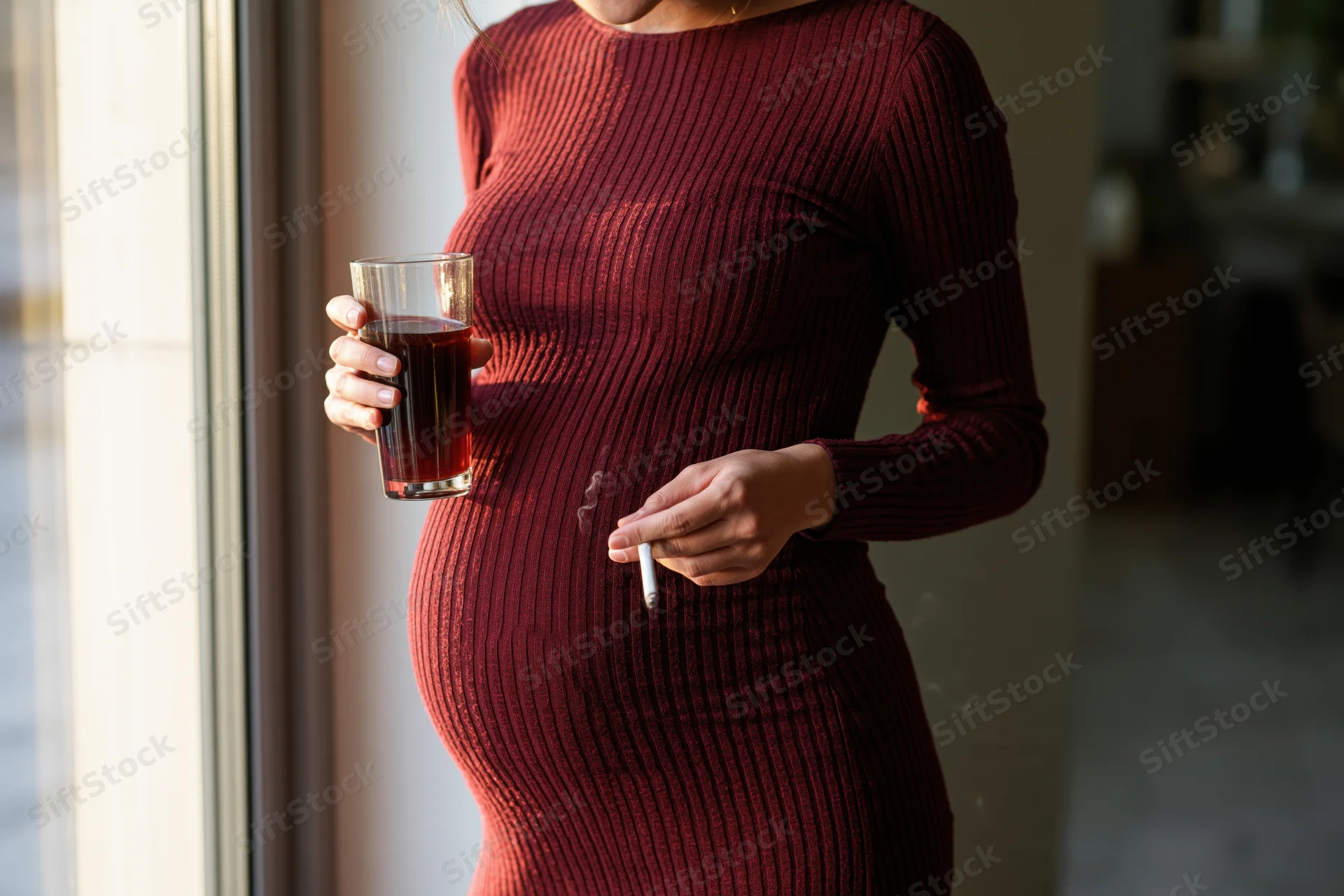 Pregnant woman holding a glass of red wine and a cigarette by a window indoors