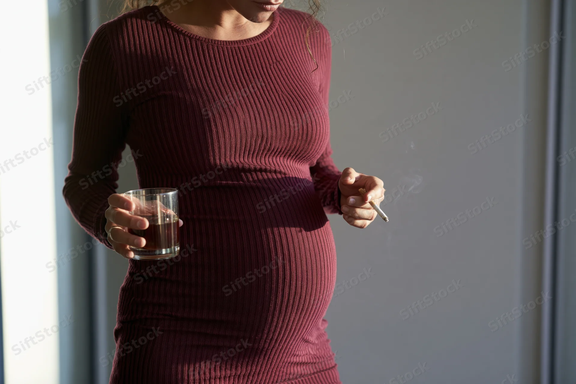 Pregnant woman holding a glass of whiskey and smoking a cigarette by a window