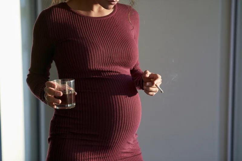 Pregnant woman holding a glass of whiskey and smoking a cigarette by a window