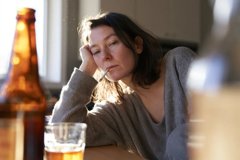 Woman smoking a cigarette while sitting at a kitchen table with a beer bottle and glass nearby