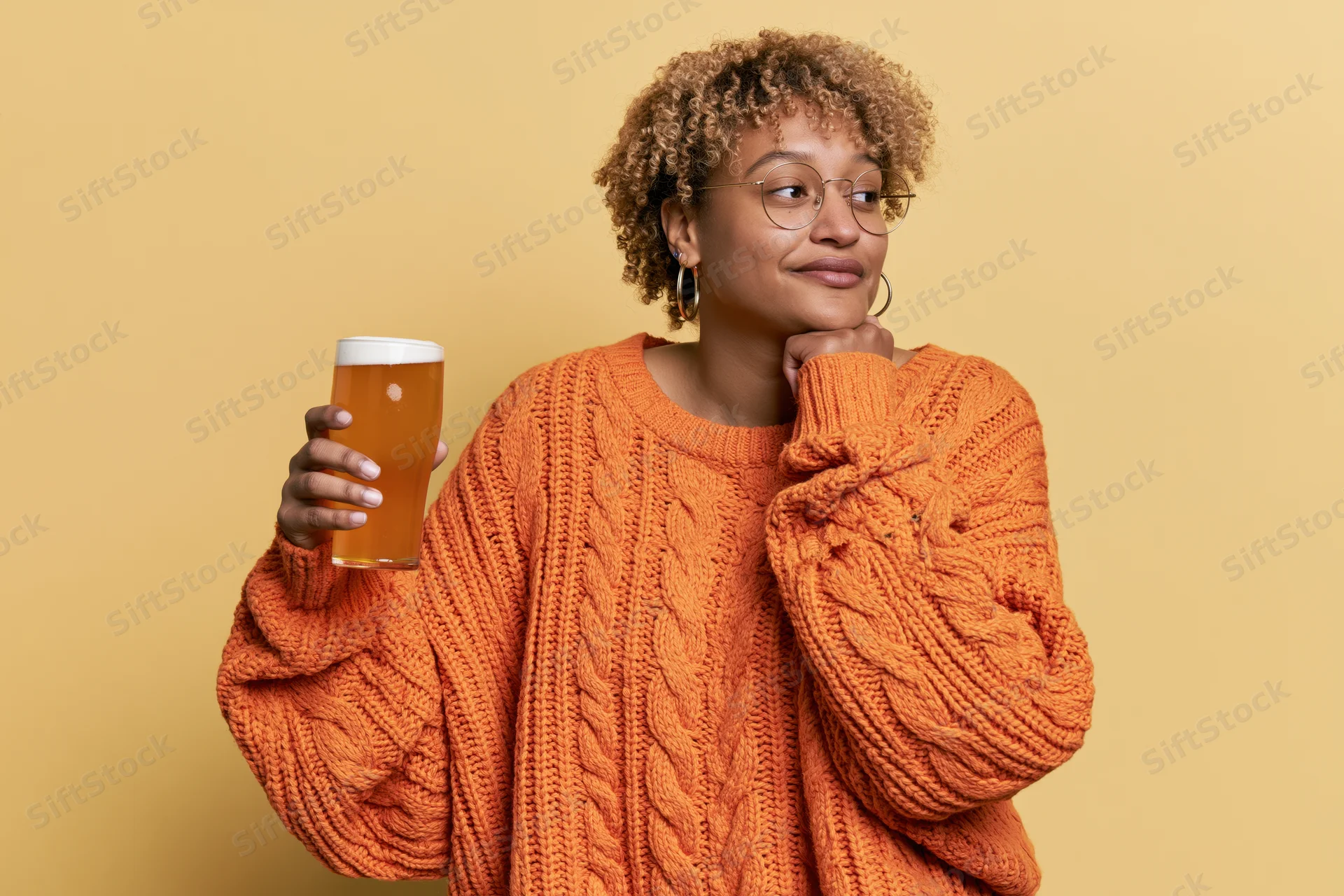Young woman in orange cableknit sweater holding beer glass, resting chin on hand, looking aside in studio setting