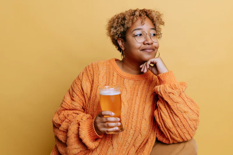 Young woman in orange knit sweater holding beer glass, smiling thoughtfully against yellow background