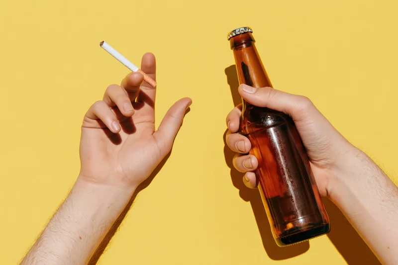 Two hands holding a cigarette and a beer bottle against a yellow background