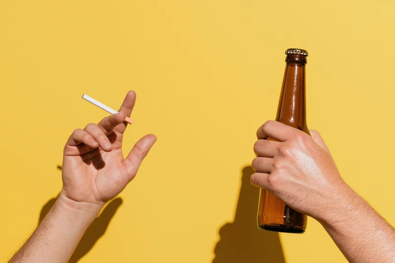 Two hands holding a cigarette and a beer bottle against a yellow background