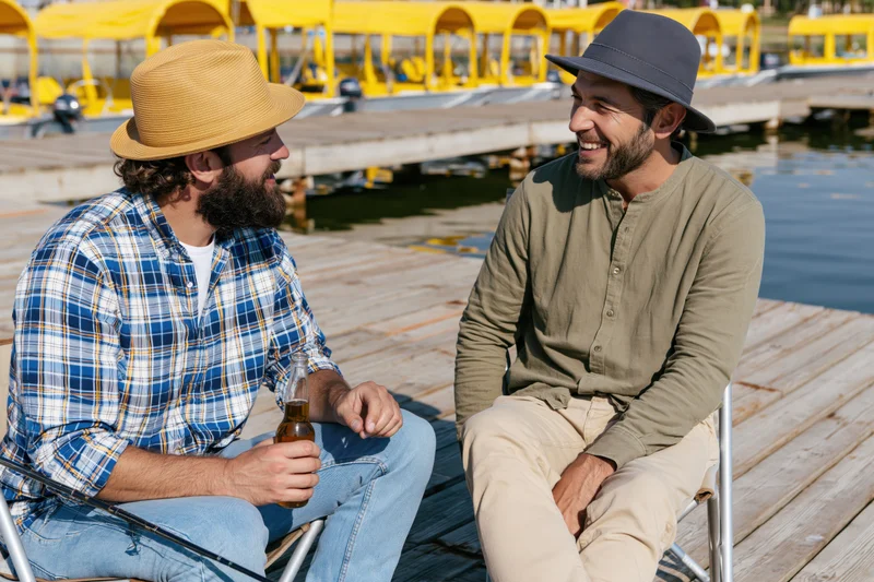 Two men sitting on a wooden dock by the water, talking and drinking beer, one holding a fishing rod, wearing hats and casual clothes