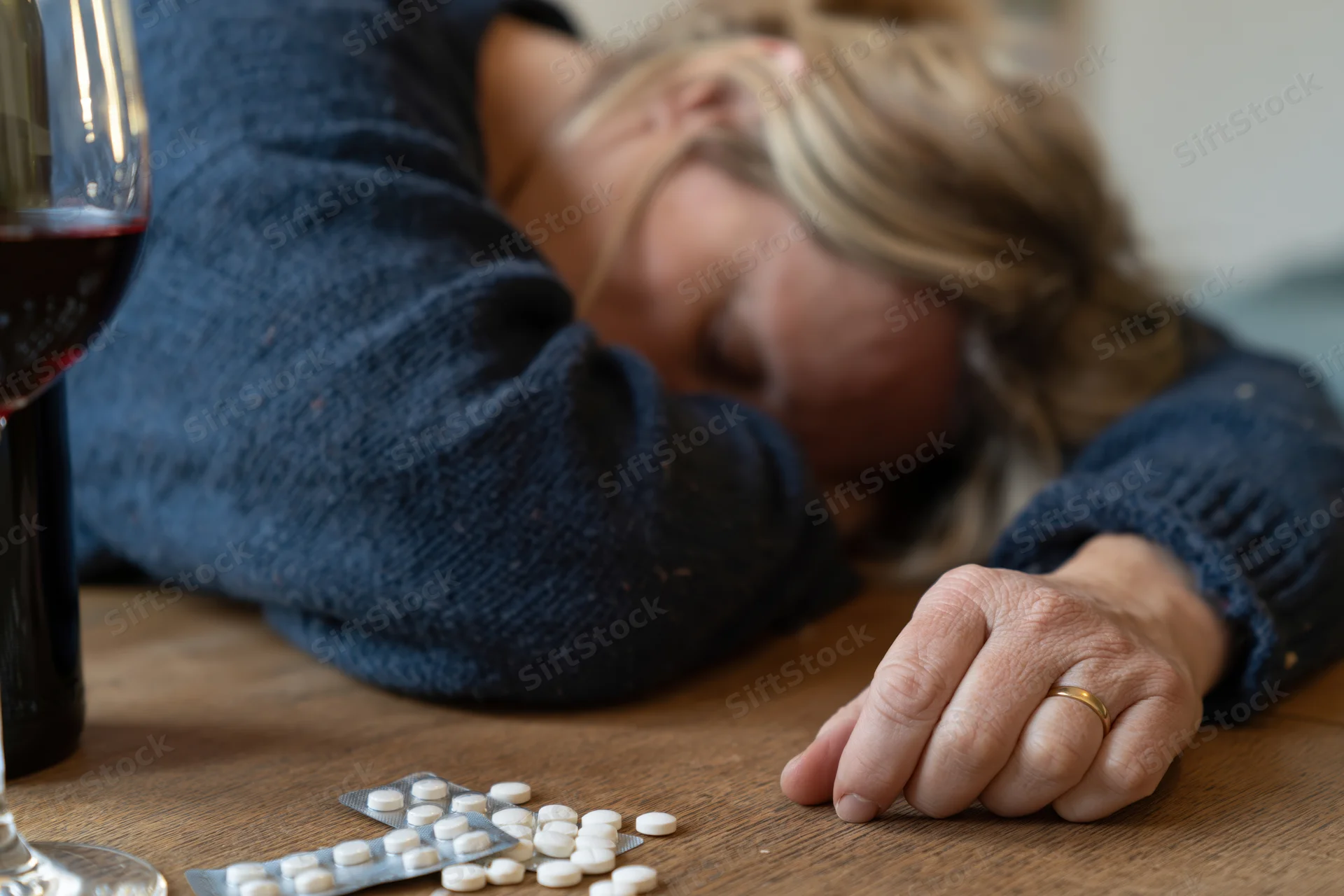 Woman sleeping with head on table beside pills and wine glass