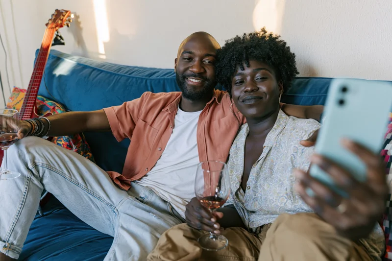Couple smiling and taking a selfie on a blue couch while holding wine glasses and a smartphone