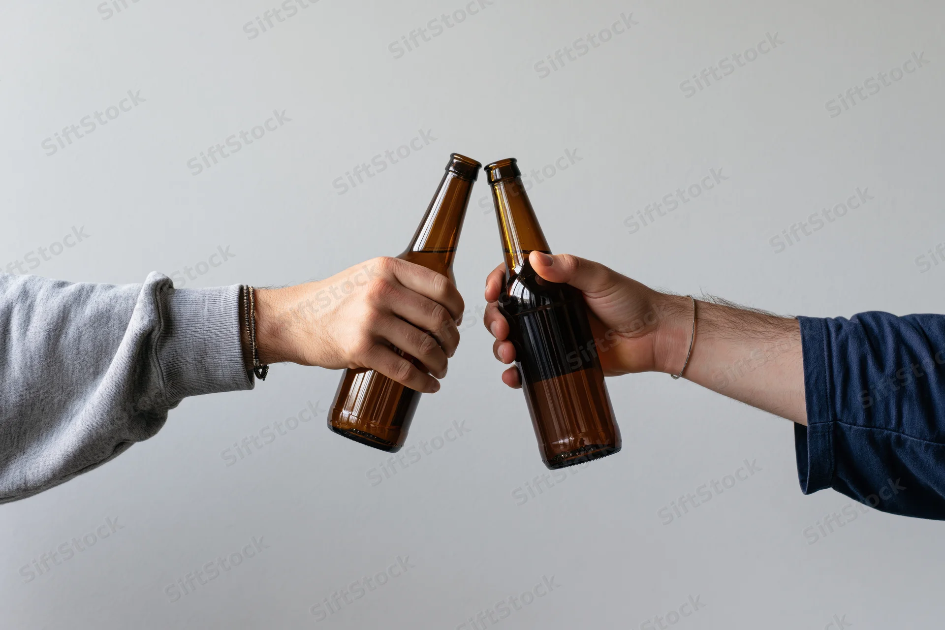 Two hands clinking brown glass beer bottles against a white background
