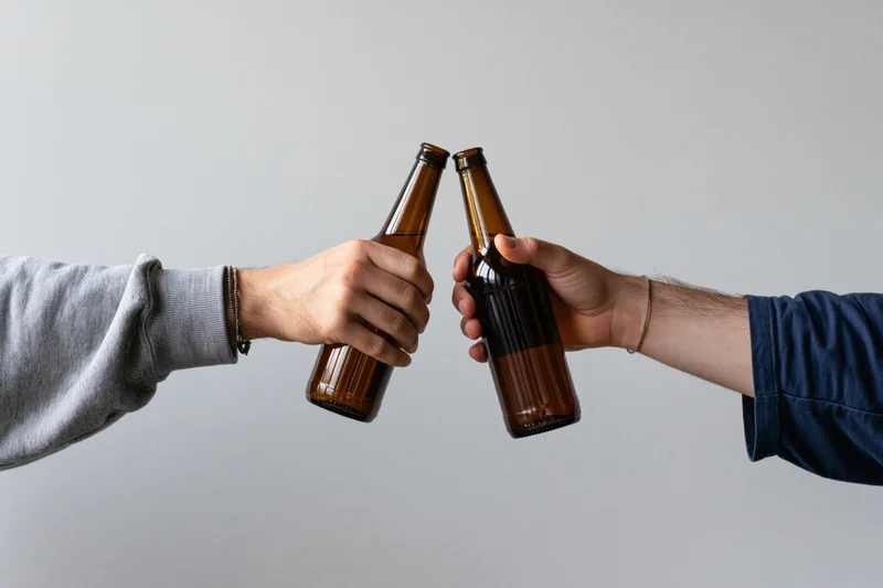 Two hands clinking brown glass beer bottles against a white background