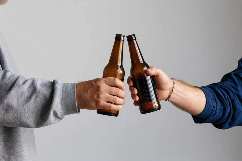 Two men clinking brown glass beer bottles in a toast, close-up view of hands and arms against a plain background