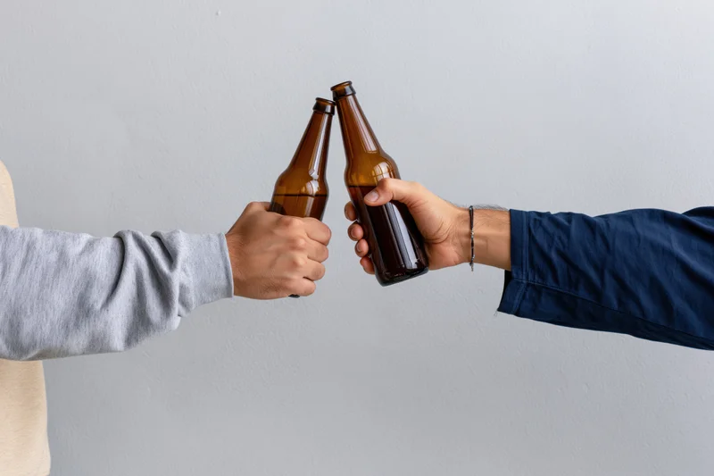 Two people clinking brown glass beer bottles in a close-up shot against a plain white background