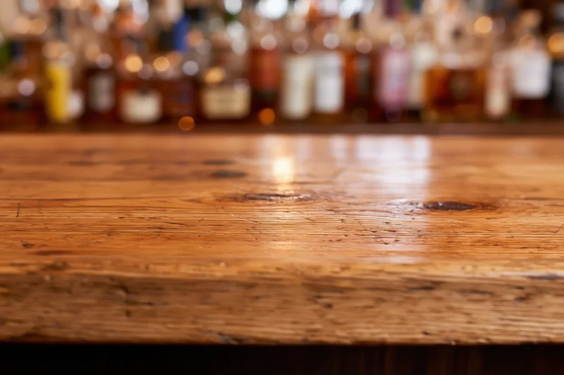 Wooden bar counter with blurred bottles in background, warm lighting, copyspace on surface