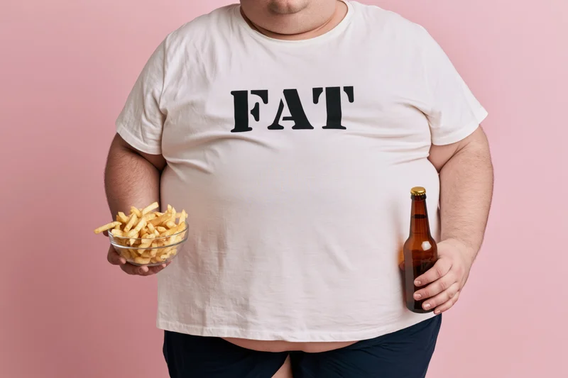 Man with large belly wearing white tshirt printed with fat holding beer bottle and bowl of fries against pink background