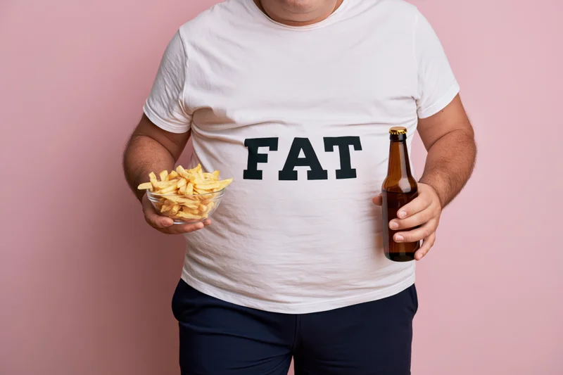 Man with belly wearing white tshirt printed with fat holding beer bottle and bowl of fries against pink background