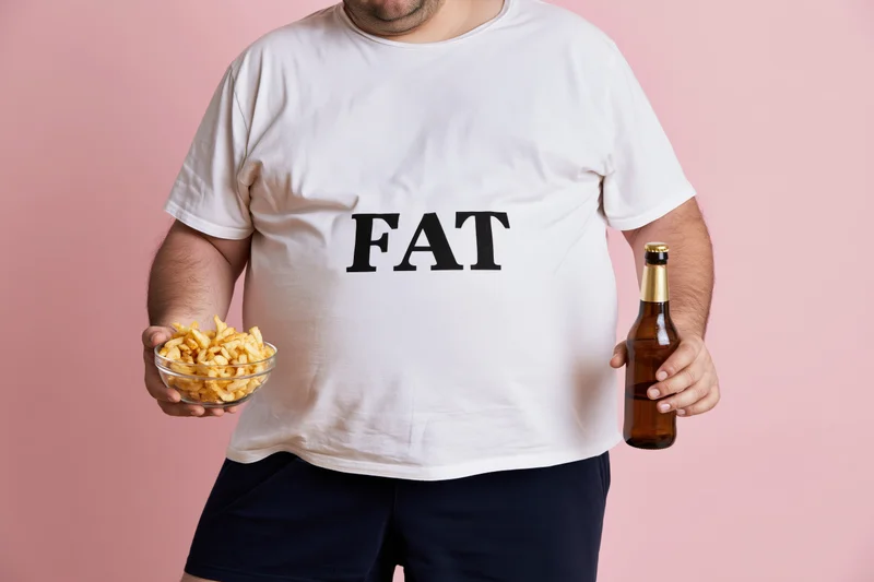 Man holding bowl of fries and beer bottle, wearing white tshirt with fat text, against pink background