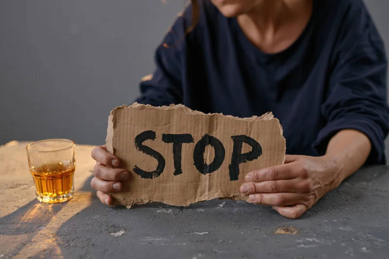 Woman holding a torn cardboard sign with stop written on it at a table beside a glass of whiskey