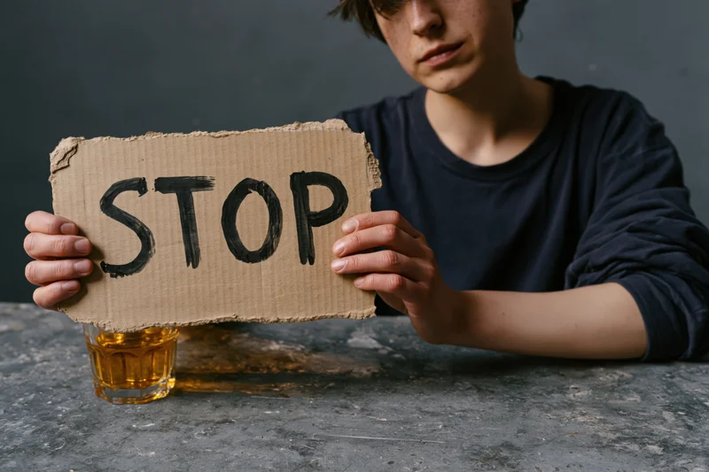 Young woman holding cardboard sign with stop written on it, sitting at table with whiskey glass beside her