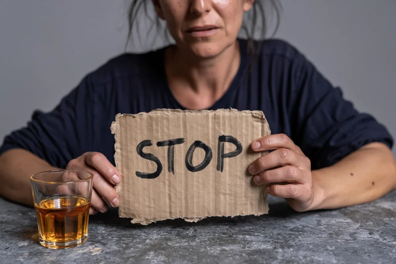 Woman holding a cardboard sign with stop written on it next to a glass of whiskey on a concrete table