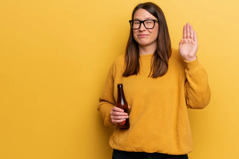 Woman holding beer bottle and raising hand in refusal against yellow background