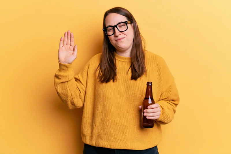 Woman holding a beer bottle and waving with one hand against a yellow background