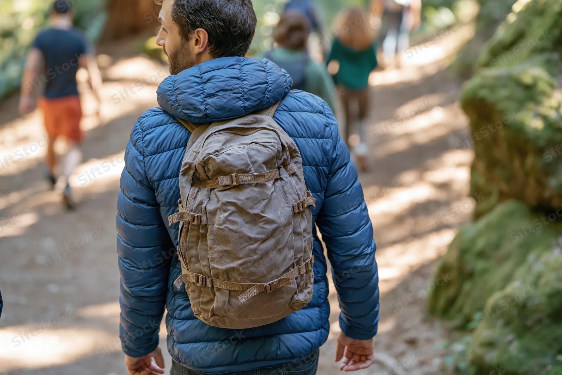 Man hiking on forest trail with backpack, surrounded by trees and mossy rocks, group walking ahead