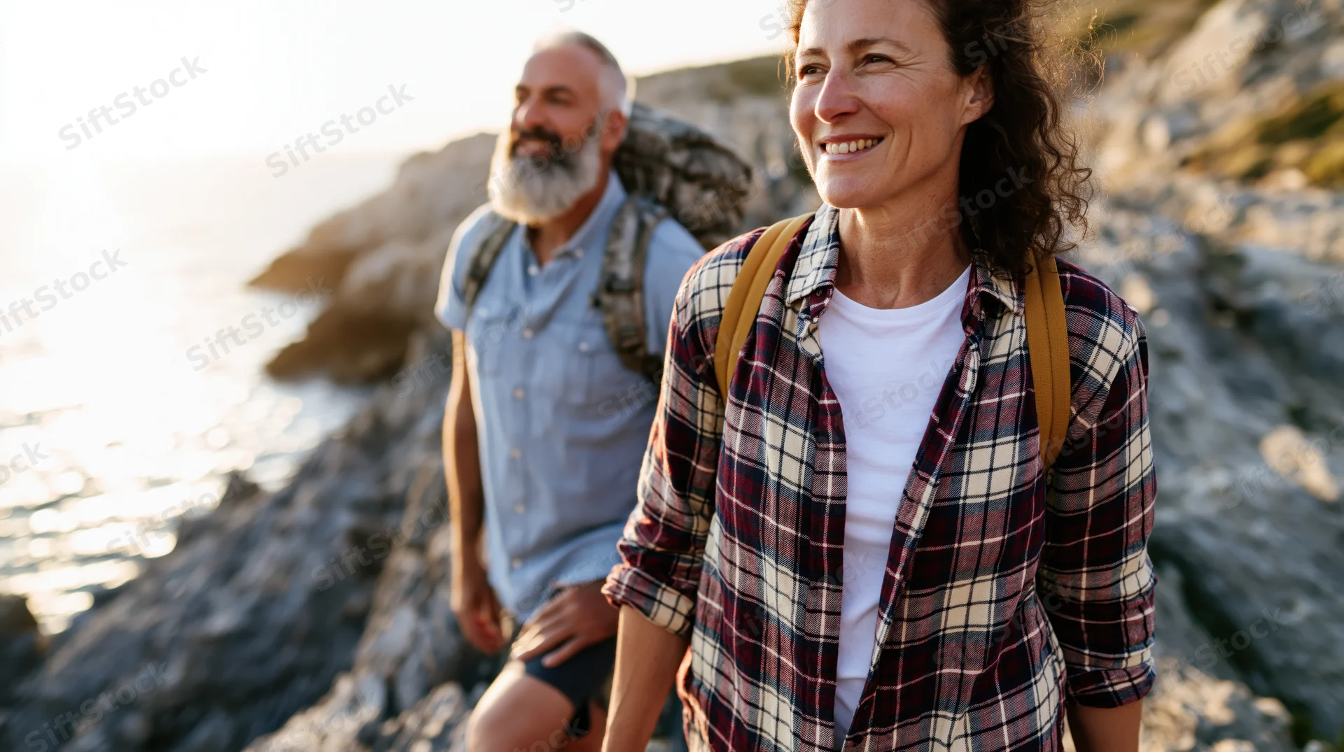 Woman and man hiking along coastal cliff trail with backpacks, smiling at sunset