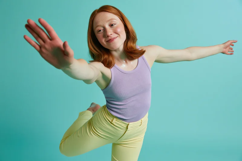 Young woman dancing in a pose with arms extended and one leg lifted against a teal background