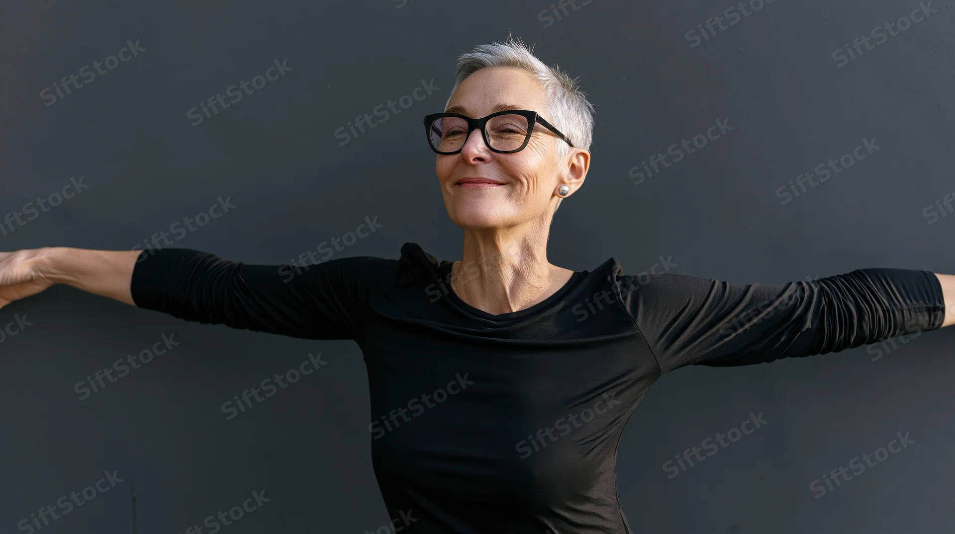 Senior woman smiling with arms outstretched against gray wall outdoors