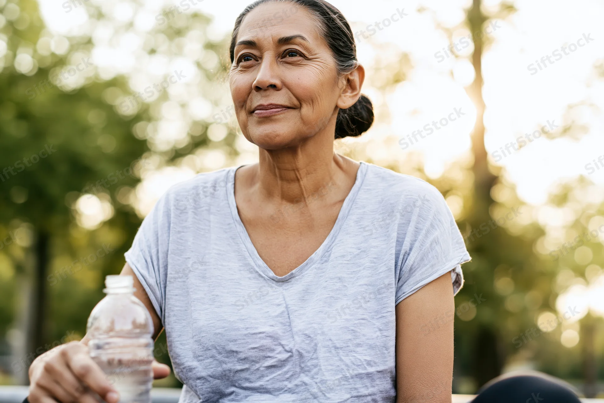 Senior woman holding water bottle outdoors in soft natural light, smiling and relaxed in a park setting