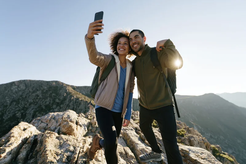 Couple taking a selfie on a mountain summit with backpacks and rocky terrain in daylight