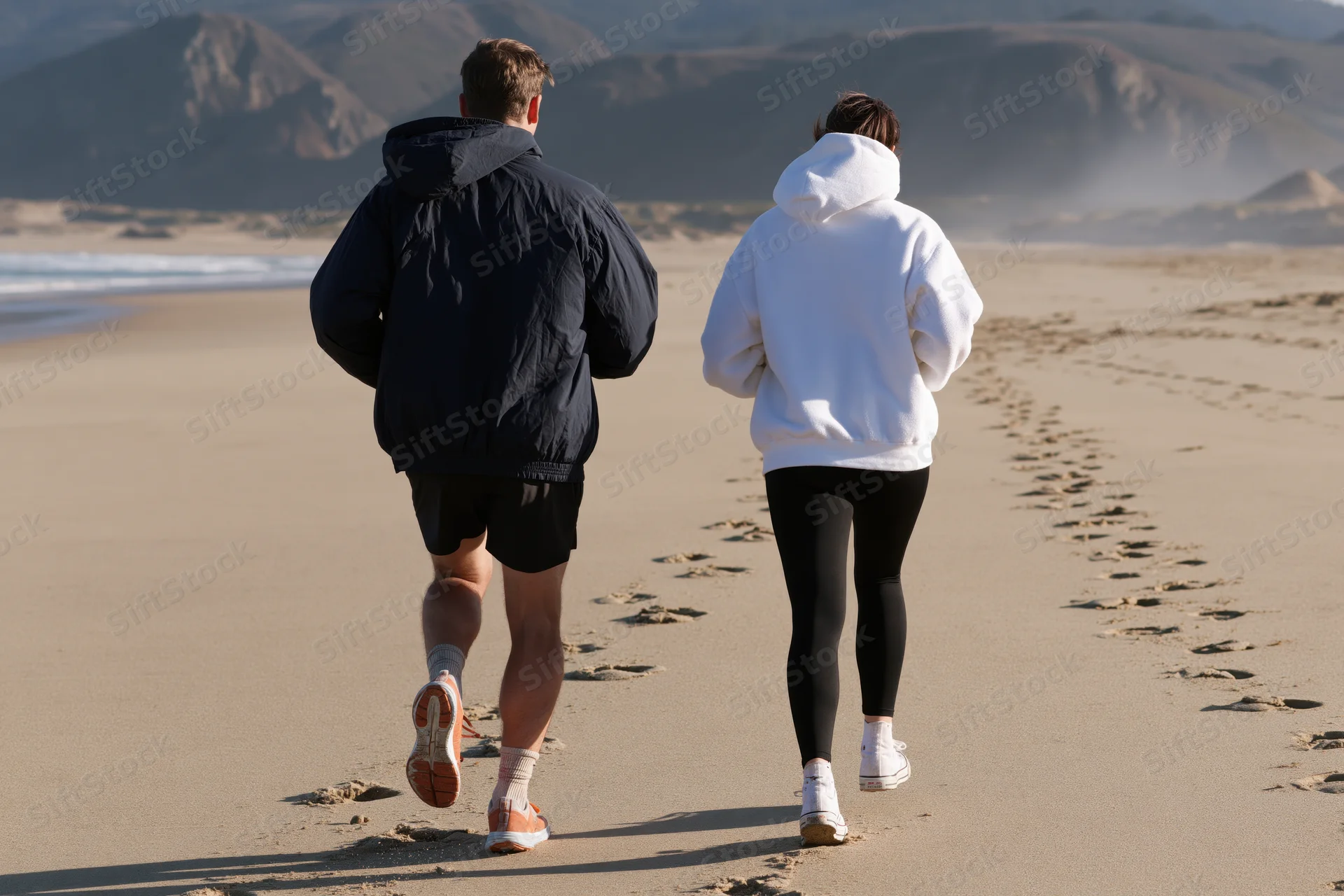 Couple running away from camera along wide sandy beach with mountains and ocean in background