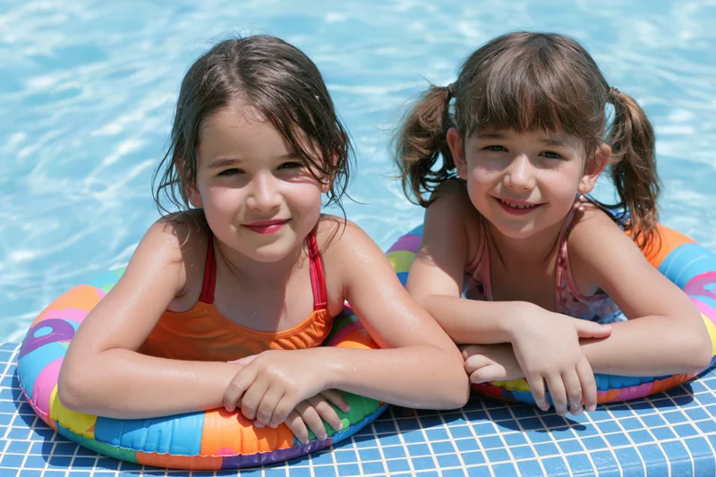 Two young girls smiling while floating on colorful inflatable rings in a bright outdoor pool