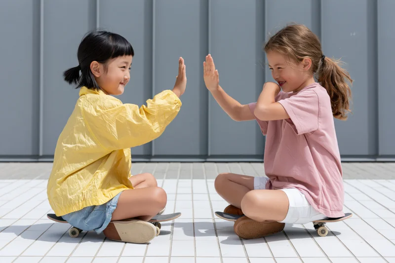 Two young girls sitting cross-legged on small skateboards outdoors, high-fiving each other on a tiled pavement