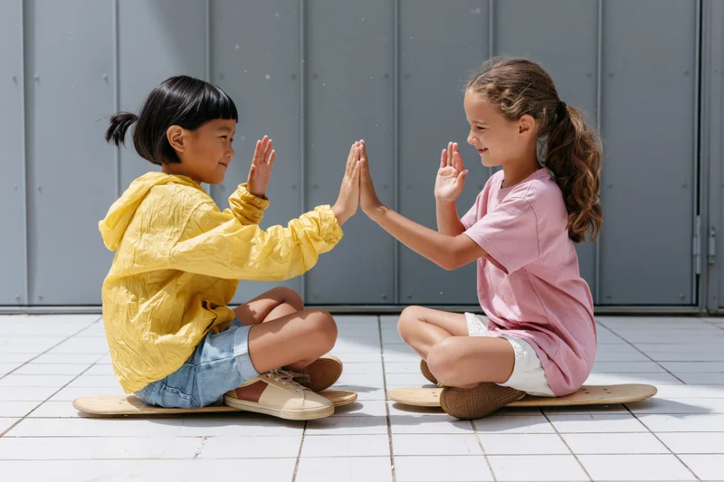 Two young girls sitting cross-legged on small skateboards outdoors, high-fiving and smiling