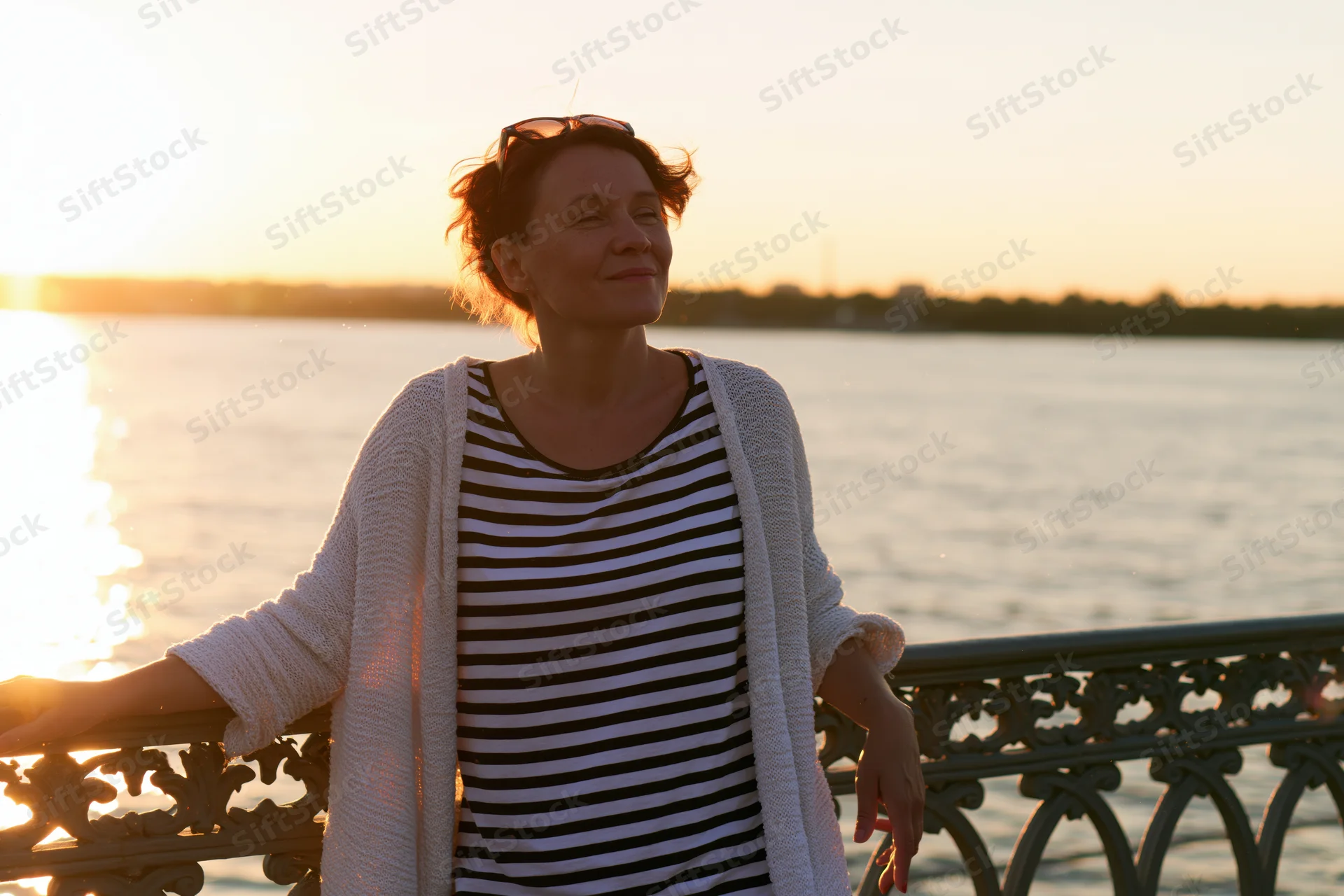 Woman leaning on decorative iron railing by waterfront at sunset