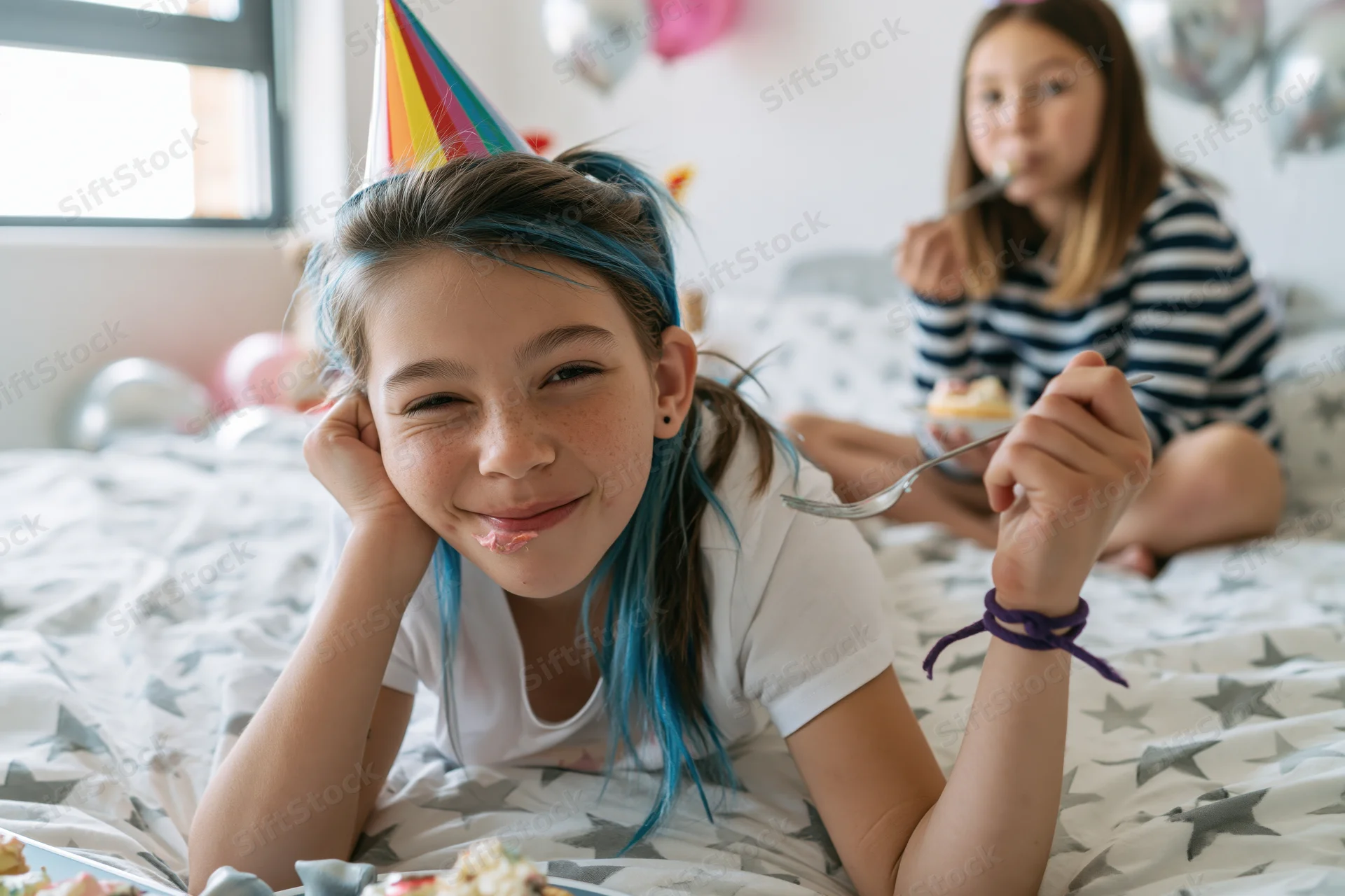 Girl winking with blue hair and party hat on bed during birthday party with cake and balloons
