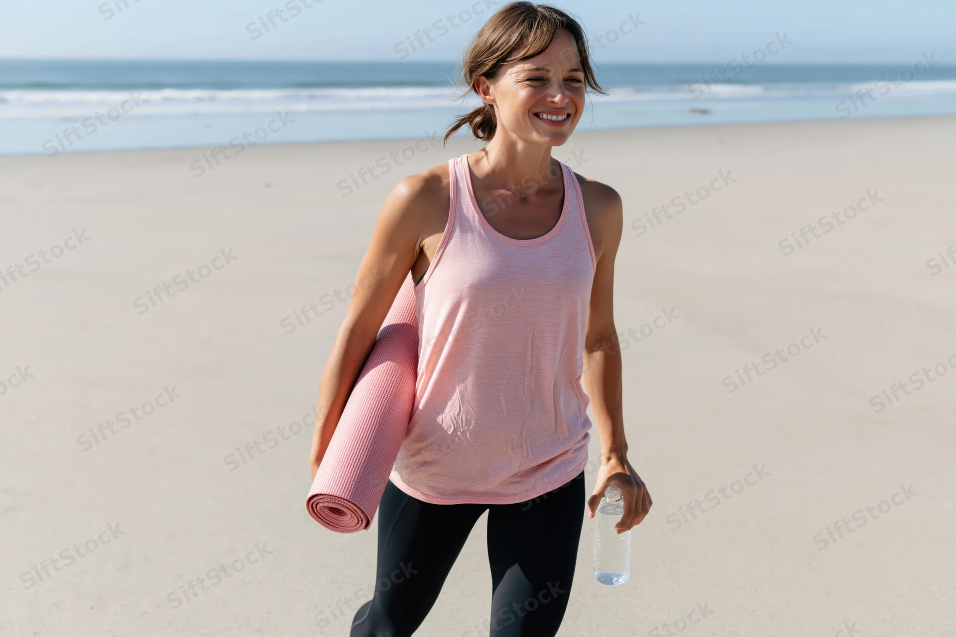 Woman walking on beach carrying yoga mat and water bottle, smiling, in athletic wear