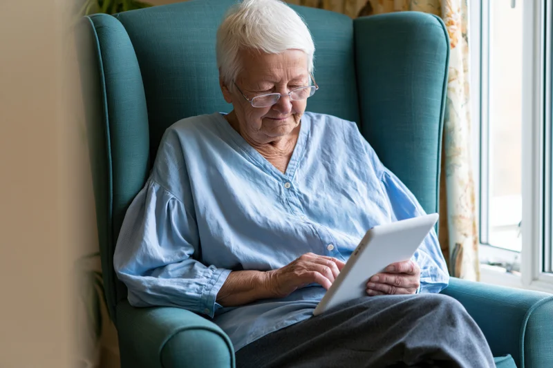Elderly woman using tablet while seated in armchair by window