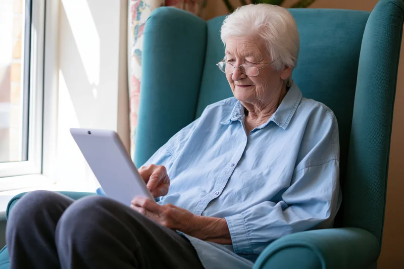 Elderly woman smiling while using tablet in armchair by window