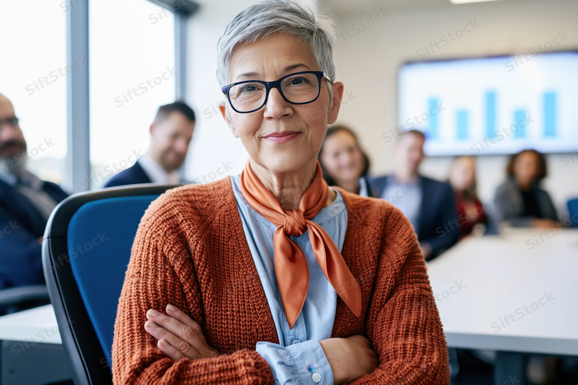 Senior woman with arms crossed smiling in office conference meeting with team and data chart on screen