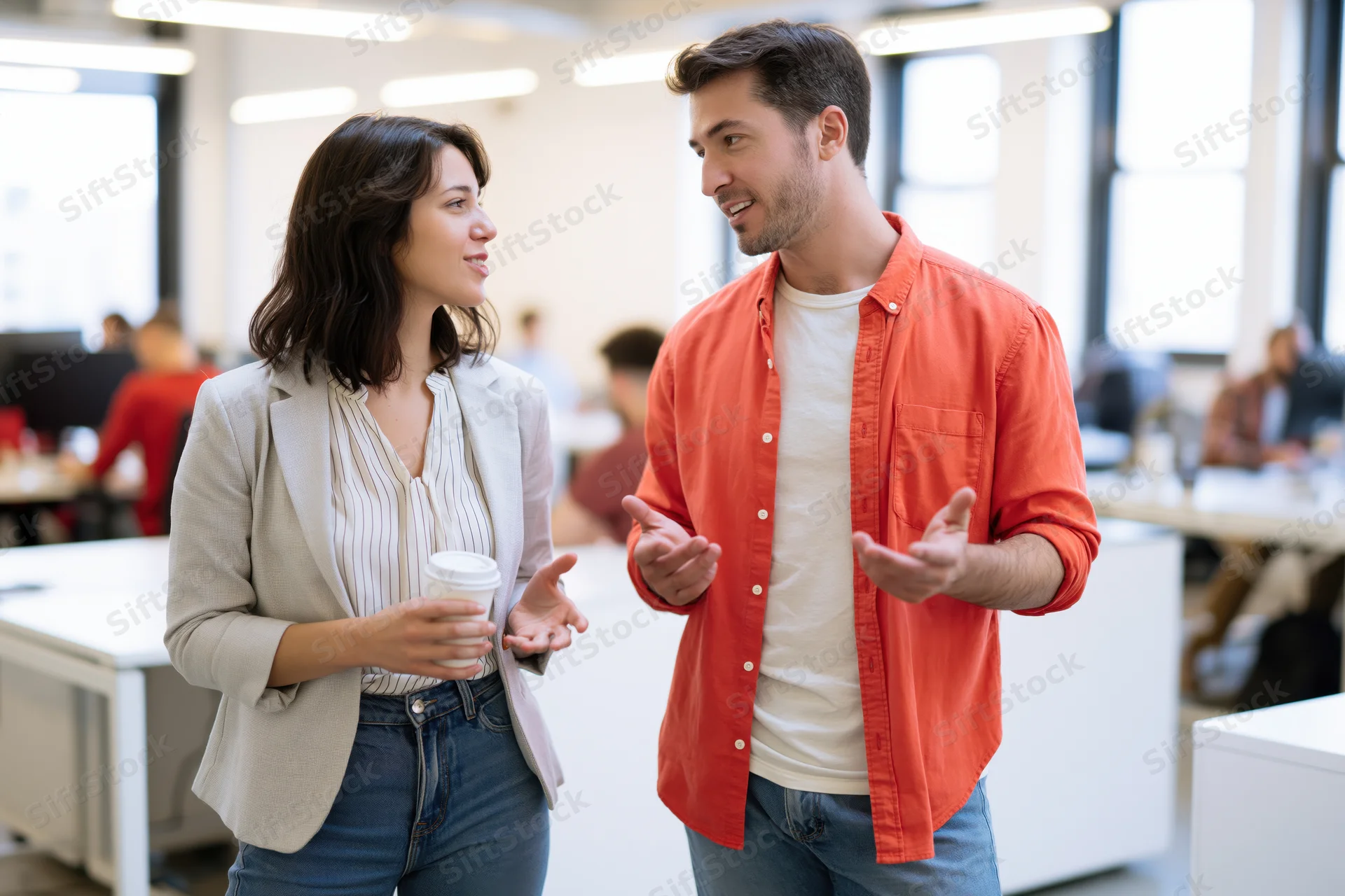 Woman and man talking and gesturing in an open-plan office with desks and windows in the background