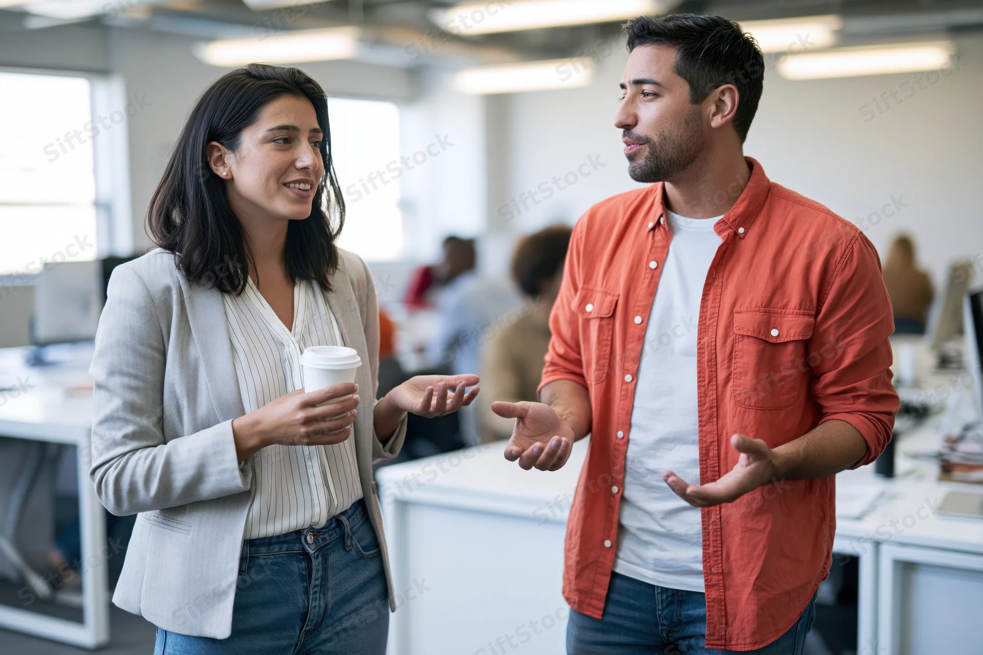 Woman and man talking and gesturing in an open office space holding coffee cups