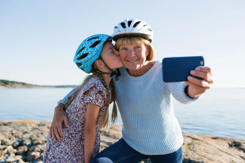 Grandmother and granddaughter taking a selfie by the water while wearing cycling helmets