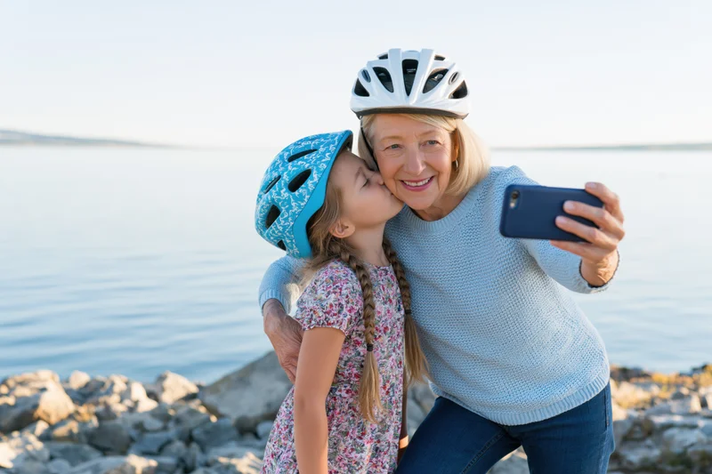 Grandmother and granddaughter taking a selfie by the lake, girl kissing her cheek, both wearing cycling helmets